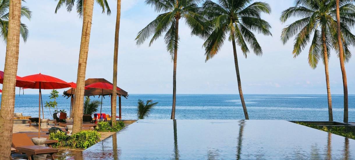 Beachfront infinity pool framed by coconut palms and red parasols, Khanom–Sichon luxury lifestyle.